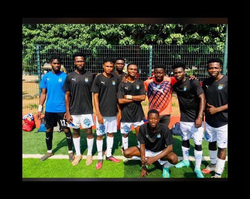 Players in black kit standing together in a group on a turf pitch