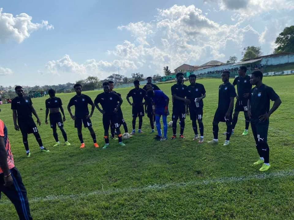 Players in black kit lined up on a grass pitch under a partly cloudy sky before kick-off