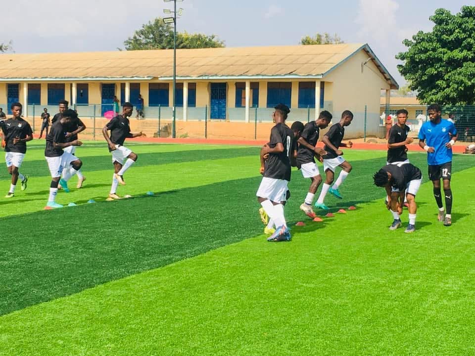 Players warming up on a turf pitch beside a training ground building