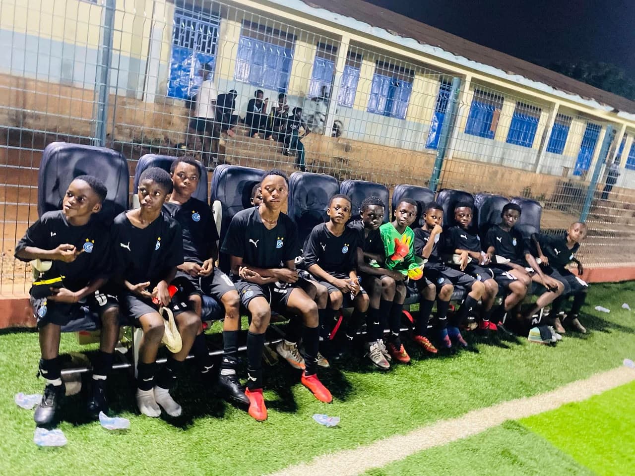 Young players in black kit seated together on the bench under floodlights