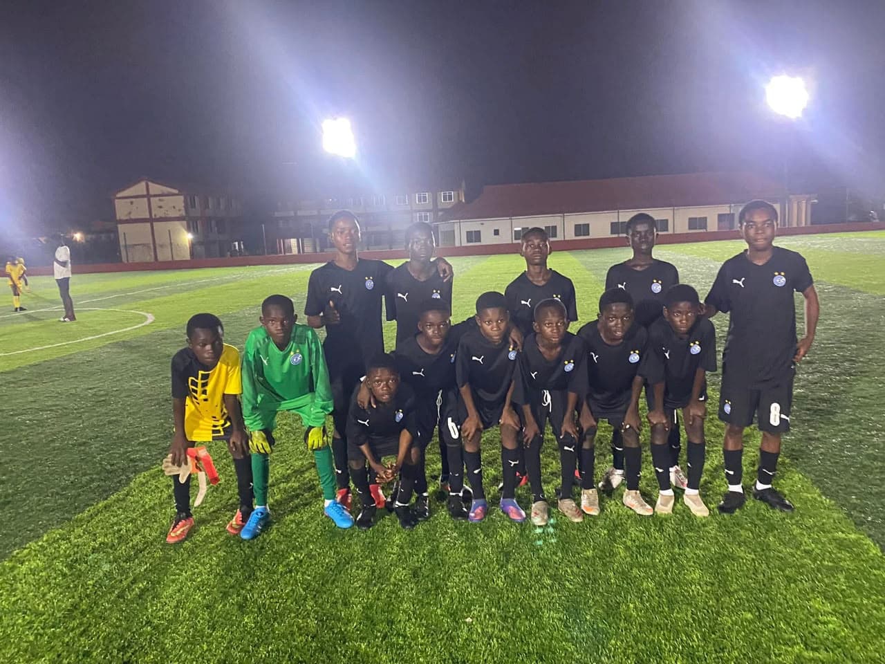 Young players and a goalkeeper in green kit posing together on the pitch at night under floodlights